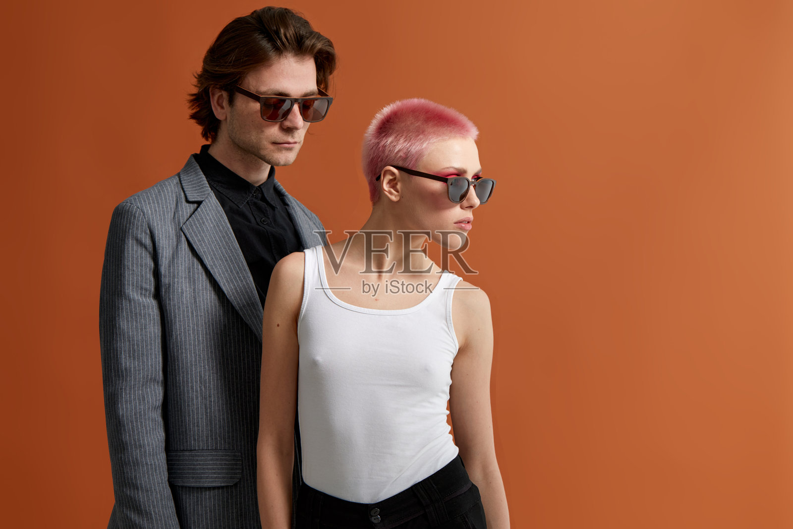 一对时髦的年轻夫妇戴着太阳镜,在棕色背景上一起摆姿势。概要肖像。照片摄影图片