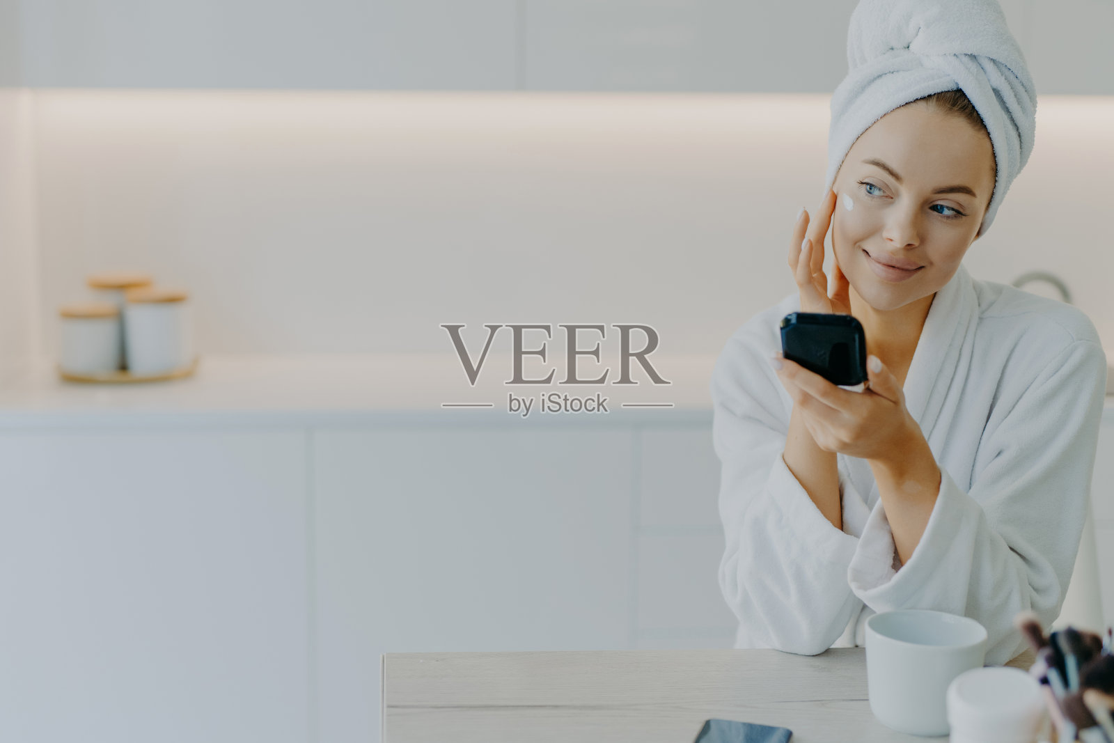 体贴美丽的年轻女子涂抹面霜,轻轻触摸脸颊,手持镜子,望向一边,穿着柔软的浴袍,对着厨房内部享受美容程序的皮肤护理照片摄影图片