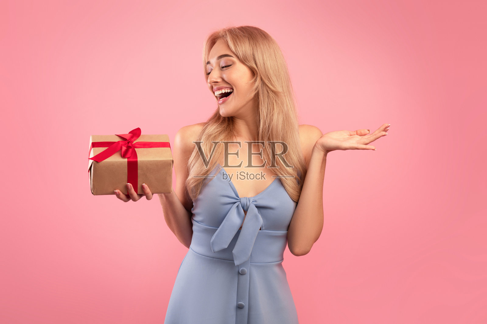 快乐的年轻女士在春天的裙子拿着包装的礼盒在粉红色的工作室背景。假日购物的概念照片摄影图片