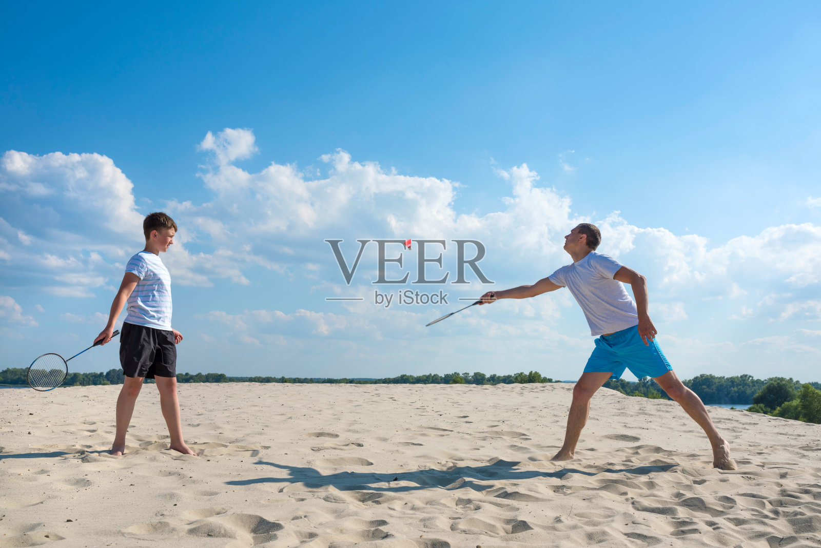 在一个阳光明媚的夏日,男孩和父亲在沙滩上打羽毛球。暑假家庭游戏。积极的休息,生活方式。照片摄影图片