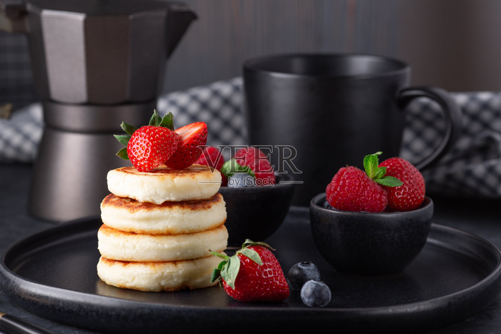 黑陶瓷盘子上的新鲜草莓和蓝莓白干酪煎饼。一杯咖啡和咖啡机照片摄影图片