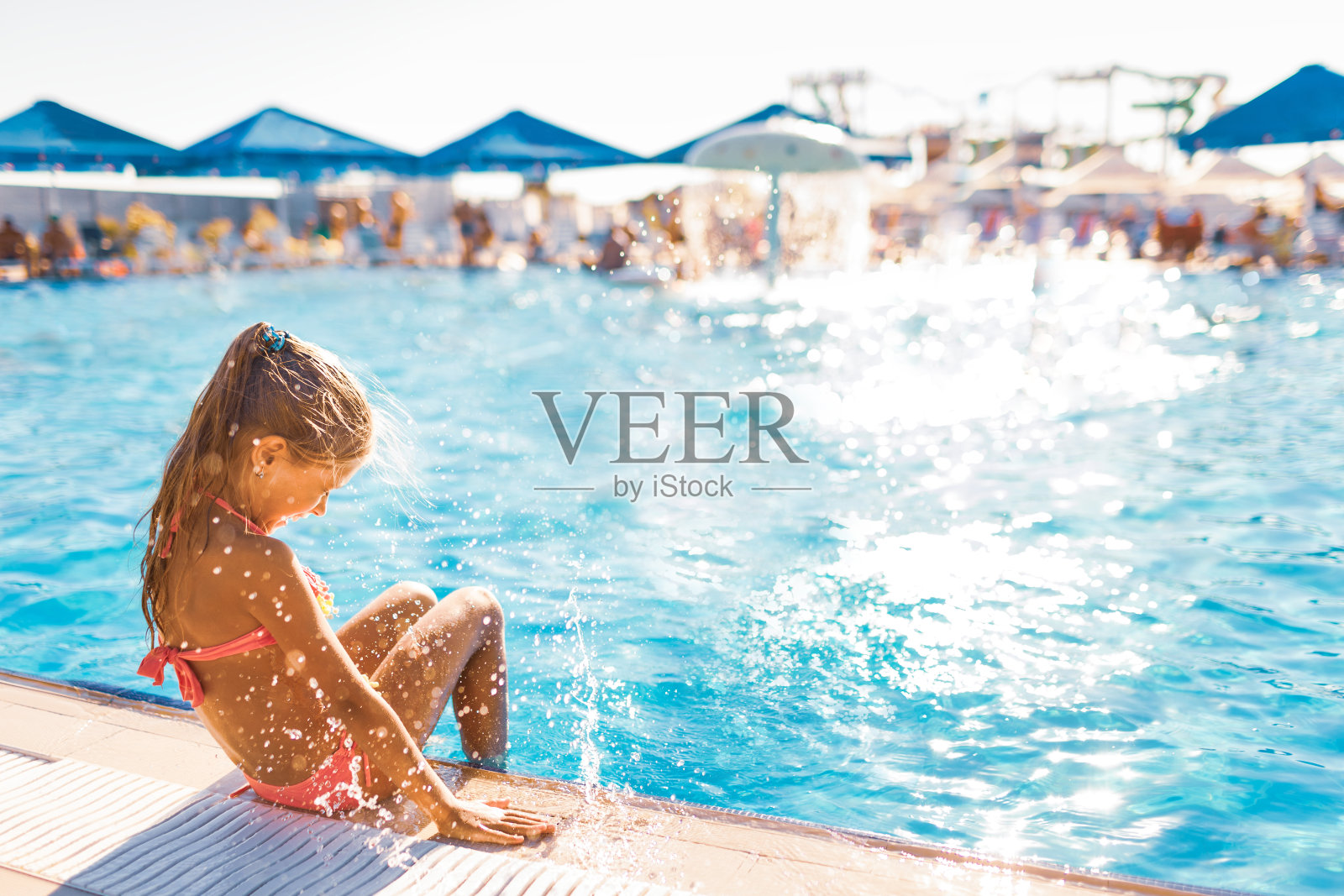 一个可爱的小女孩坐在游泳池的一边,她的脚在水里享受着阳光照片摄影图片