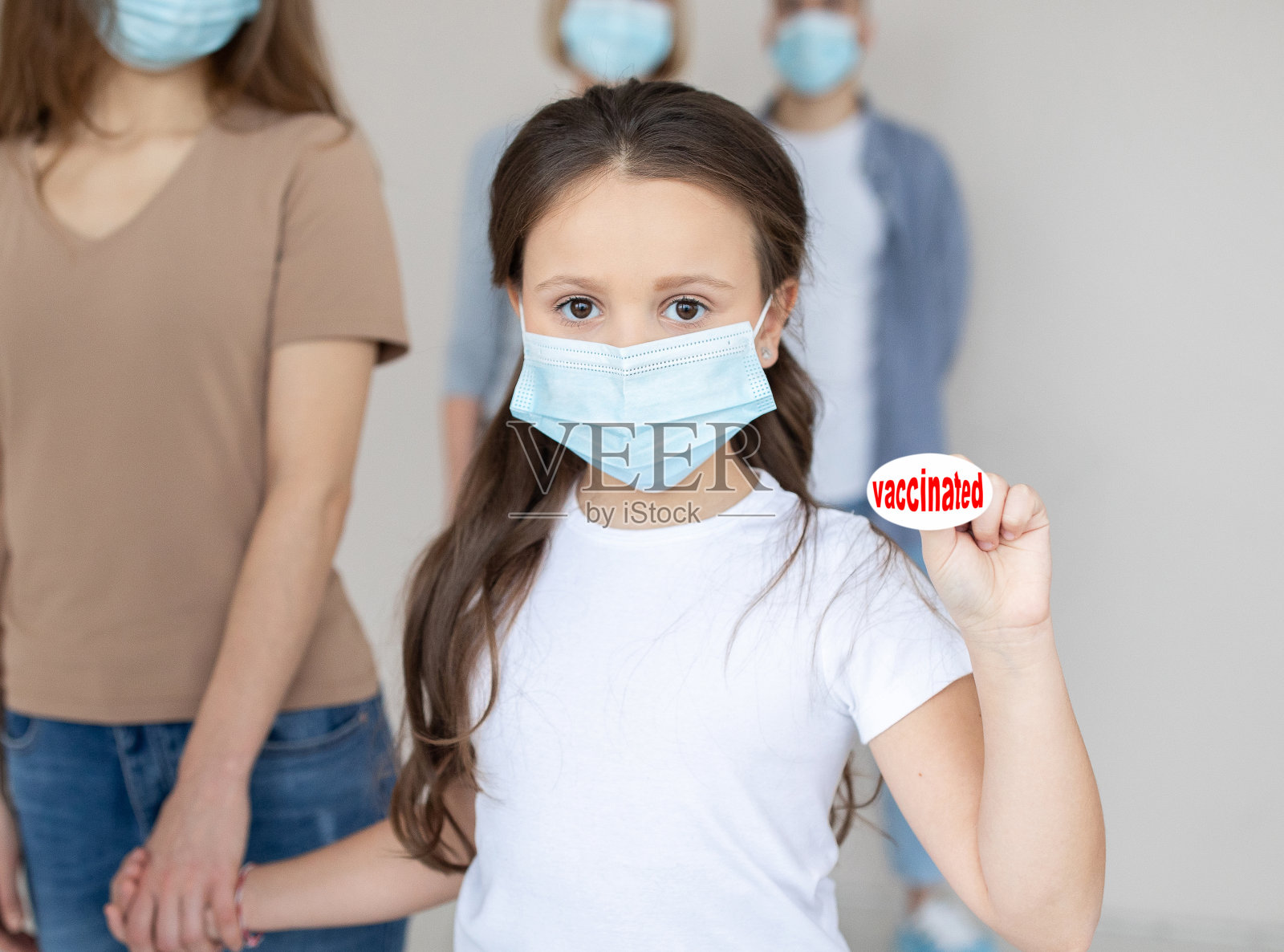 可爱的小女孩戴着口罩,牵着妈妈的手,展示接种疫苗的徽章,在医院排队照片摄影图片