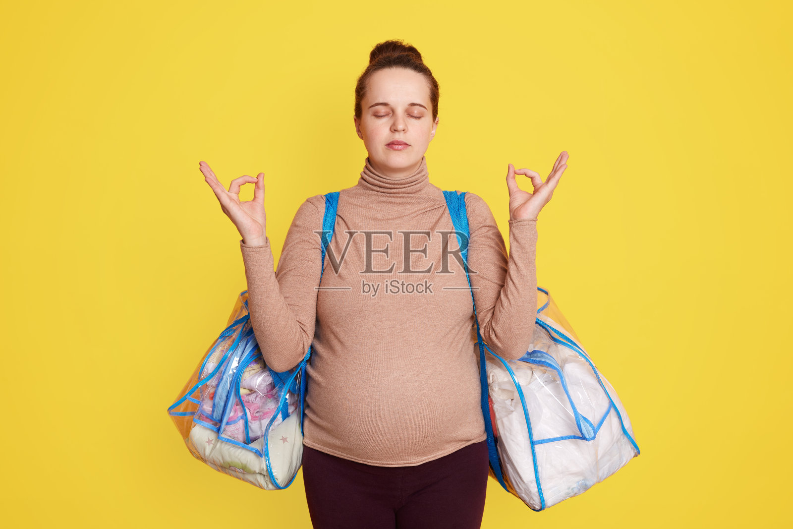 可爱冷静可爱美丽的准妈妈穿着休闲装,梳着发髻,背着妇产医院黄色的墙摆姿势,试着在分娩前放松一下,不要担心。照片摄影图片