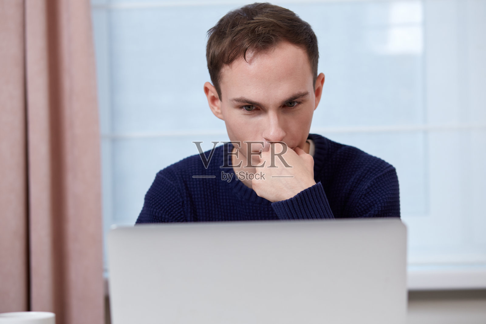 一脸严肃的年轻人在家工作。桌子上有一台笔记本电脑。照片摄影图片