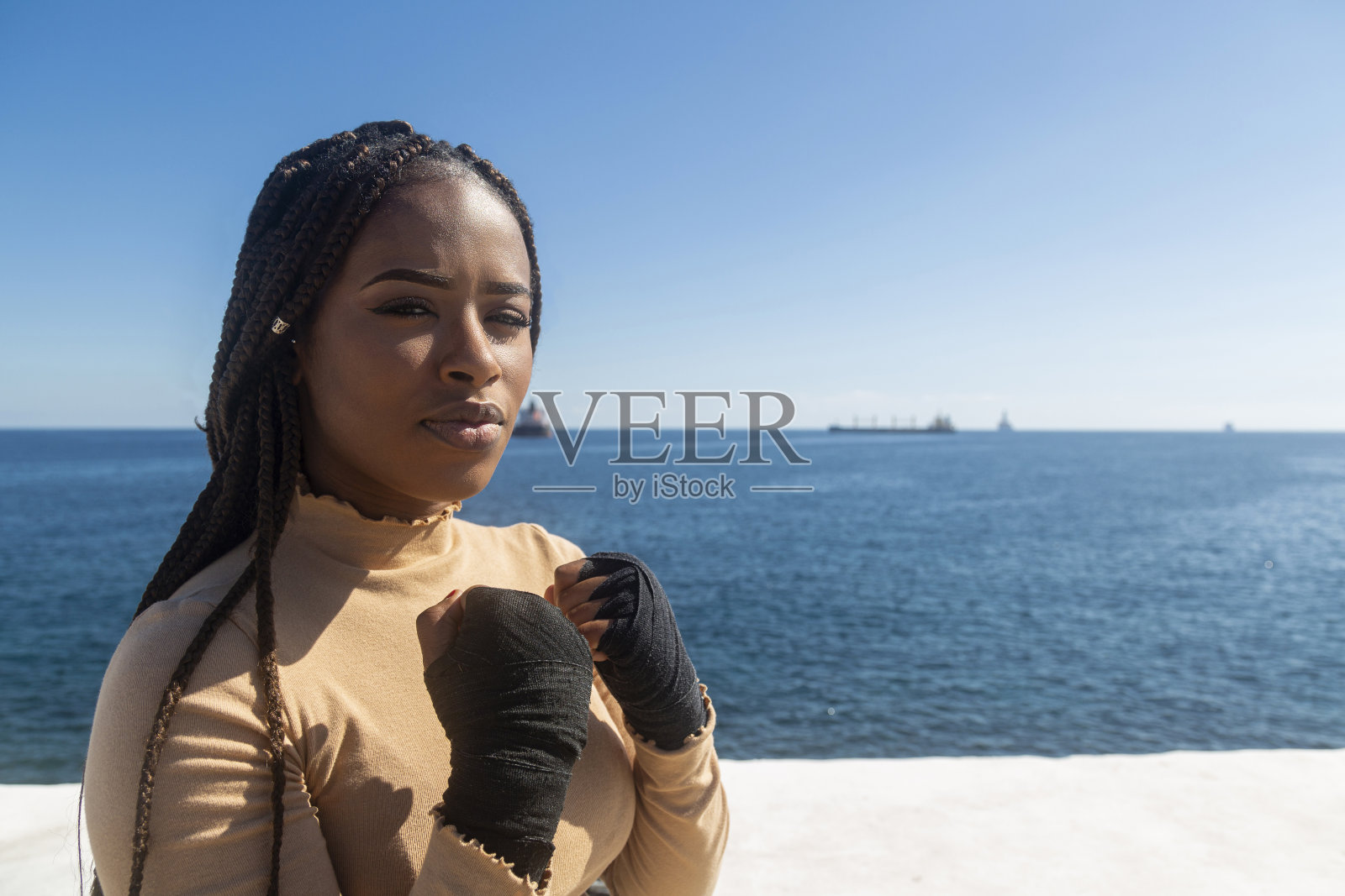 美丽年轻的美国黑人妇女看着镜头,用绷带在她的手上练习武术,拳击。天空和海洋背景,海滨。照片摄影图片