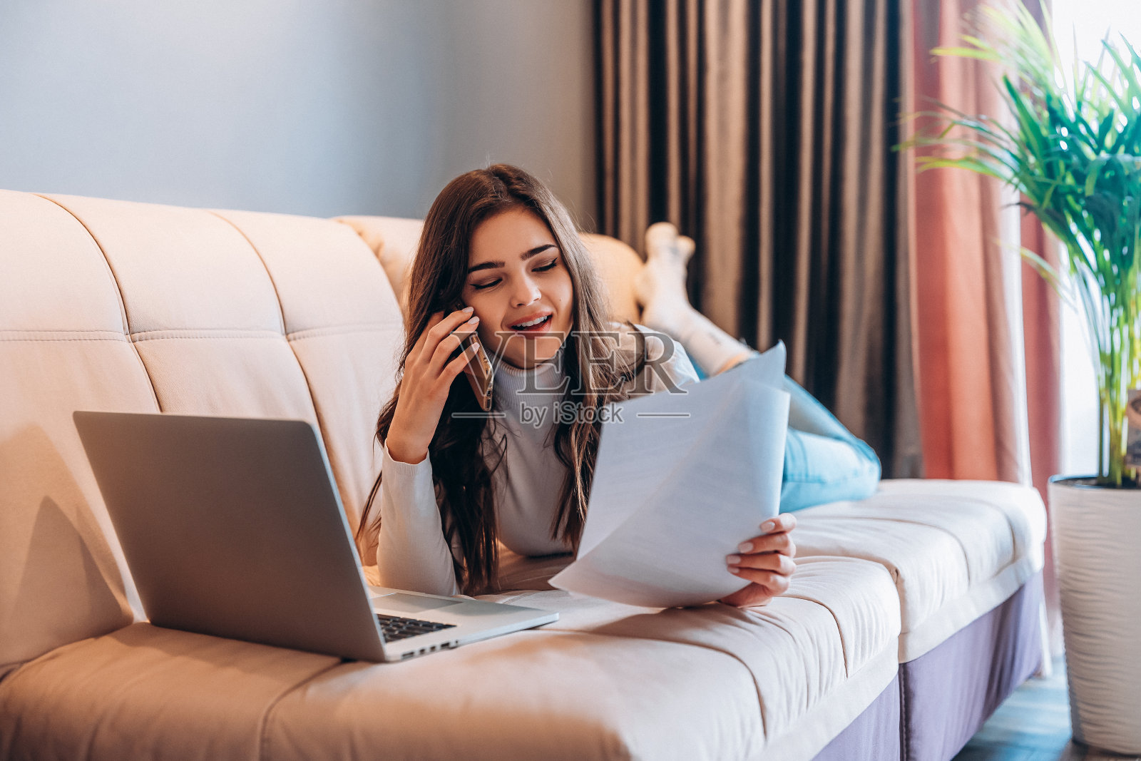 自由职业学生年轻女性坐在家里使用笔记本电脑。年轻女子躺在沙发上用笔记本电脑上网工作。漂亮的女人一边学习一边工作一边打电话照片摄影图片