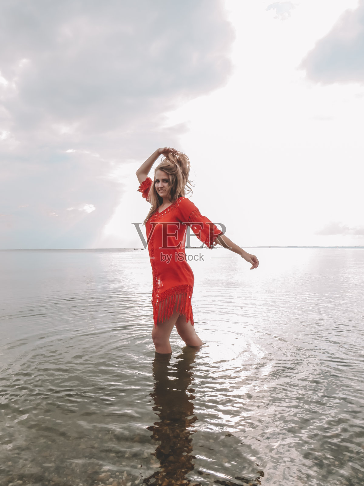 一位漂亮的年轻女子,一头金色长发,穿着红色沙滩裙,身材苗条。她走在海湾浅滩的水面上照片摄影图片