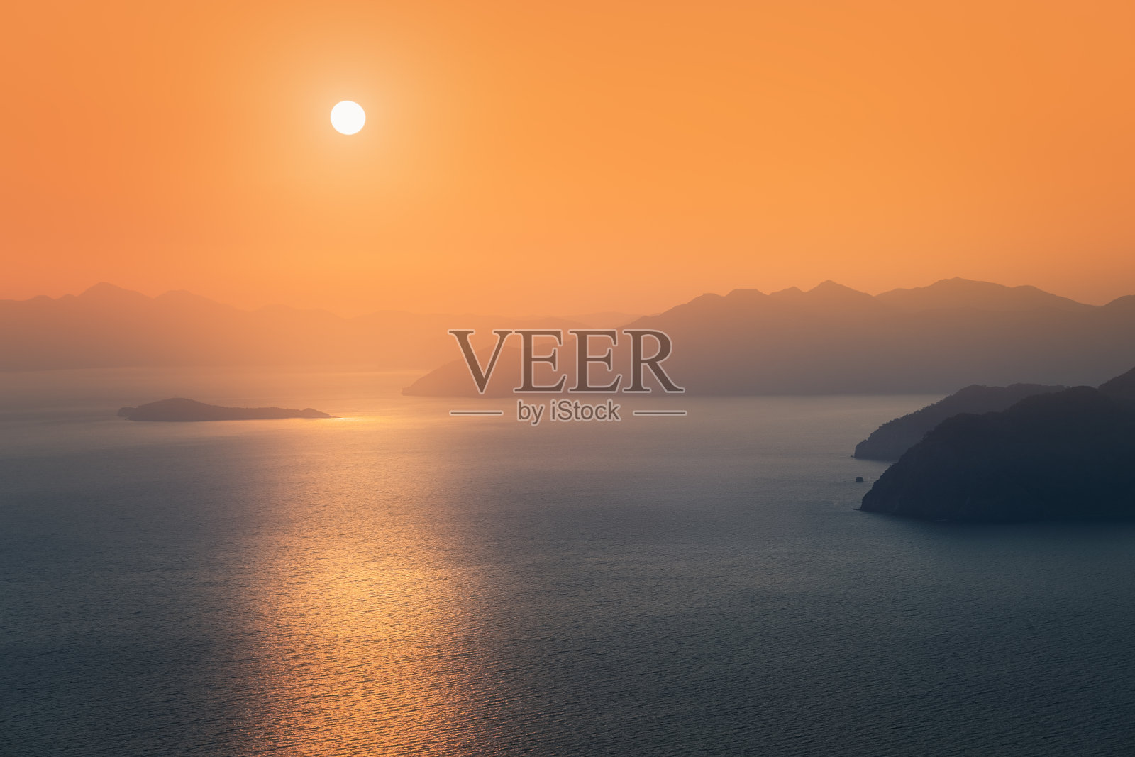 阴沉的日落,远处的太阳落在地平线上蓝色山脉的剪影后面,被地中海的海水冲刷着照片摄影图片