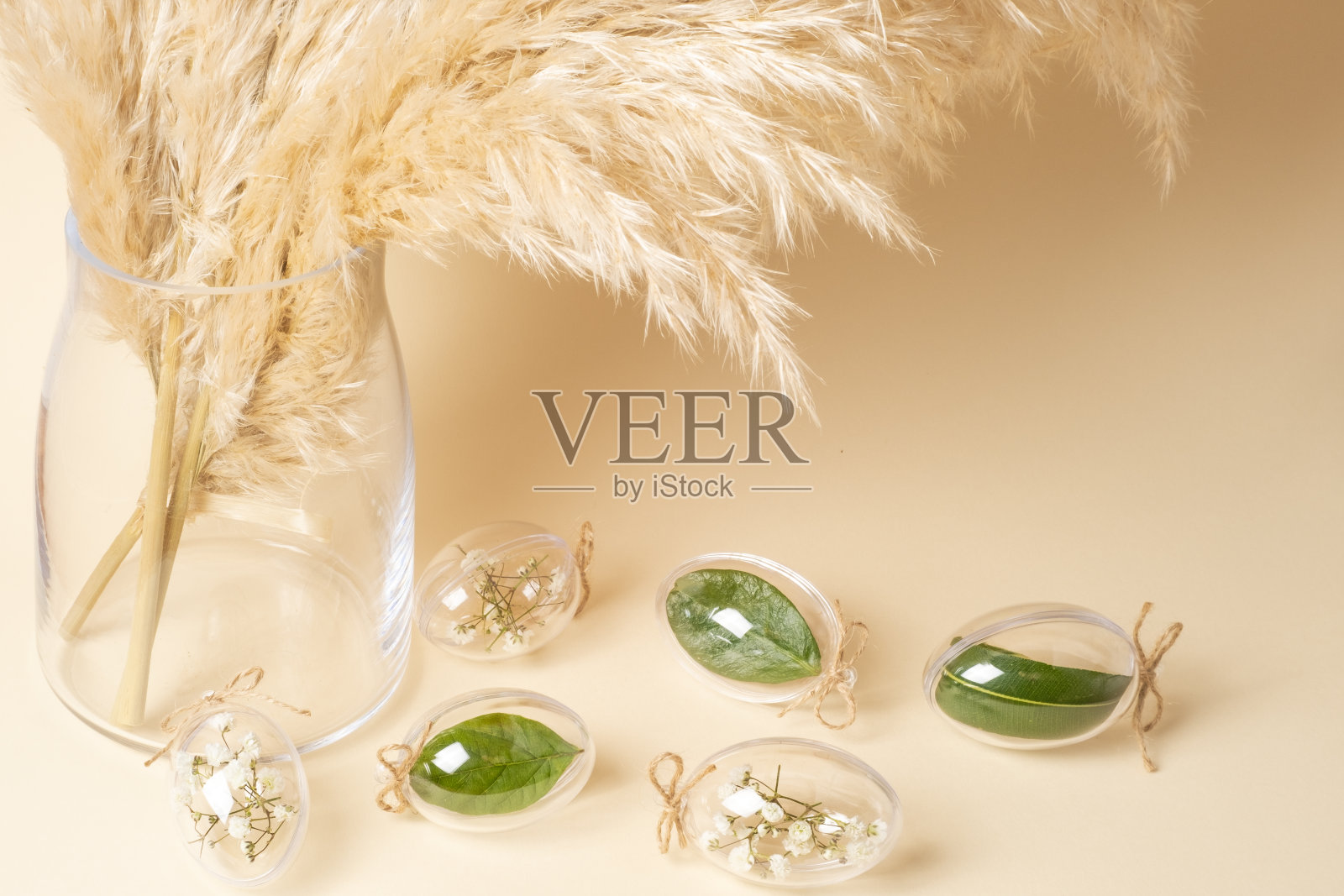 玻璃花瓶中的潘帕斯草,透明的玻璃蛋,白色的花和绿色的叶子在米色的背景。复活节假期,现代简约构图。生态时尚装饰概念。照片摄影图片