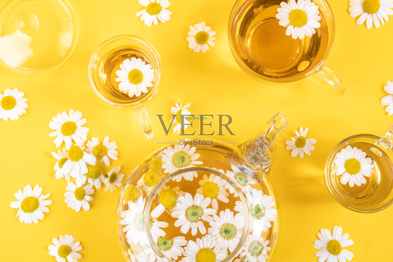 三杯茶和一个透明茶壶,黄色的背景上有洋甘菊花。甘菊茶有益健康。平铺照片摄影图片