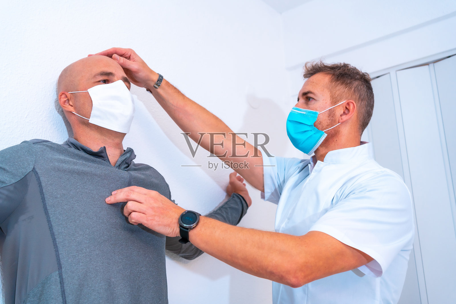 带面罩的物理治疗师用一根头发在墙上拉病人的脖子。2019冠状病毒病(COVID-19)大流行中的物理治疗和防护措施。整骨疗法、体育quiromassage照片摄影图片