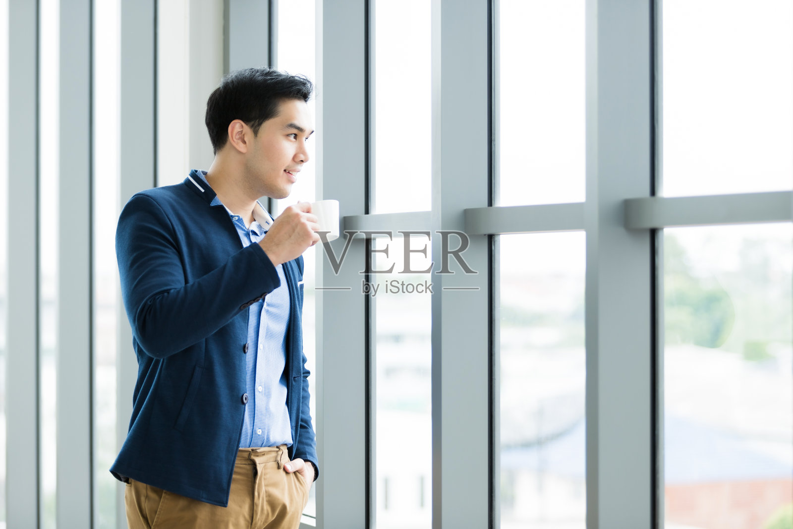 快乐心情的肖像亚洲年轻商人工作拿着一个咖啡杯穿西装的男人在蓝色夹克和蓝色衬衫看着窗户在办公室的房间背景照片摄影图片