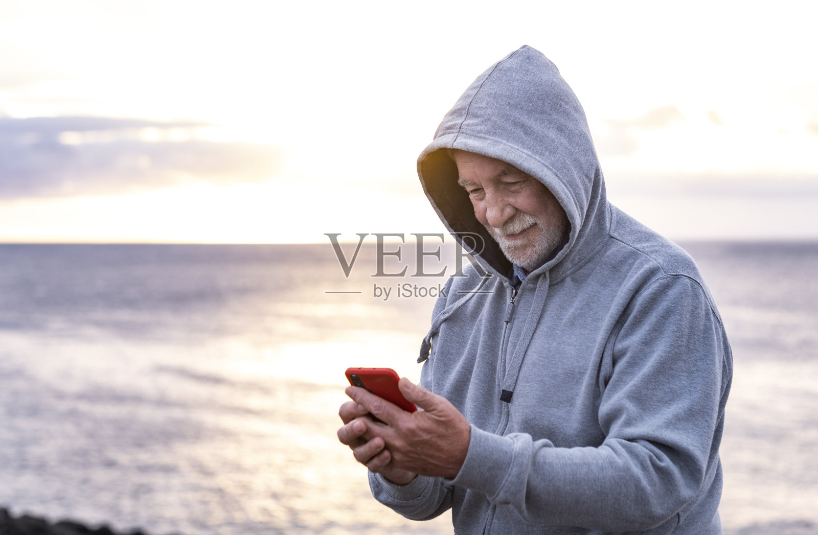 一位面带微笑的老人戴着兜帽在海上享受寒冷的冬日。在夕阳西下时看手机照片摄影图片