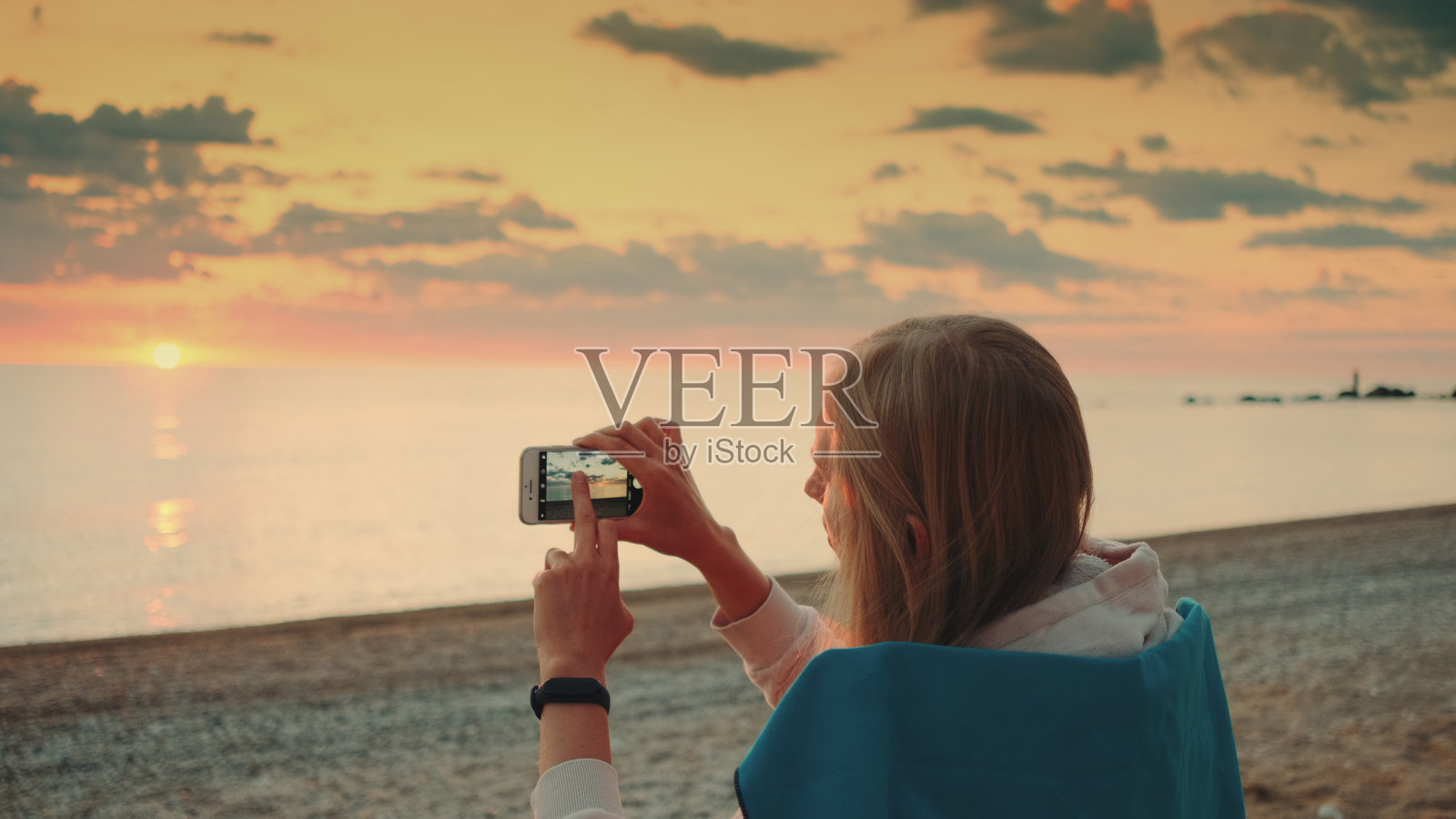 年轻女子坐在海边用智能手机拍摄日落的视频照片摄影图片