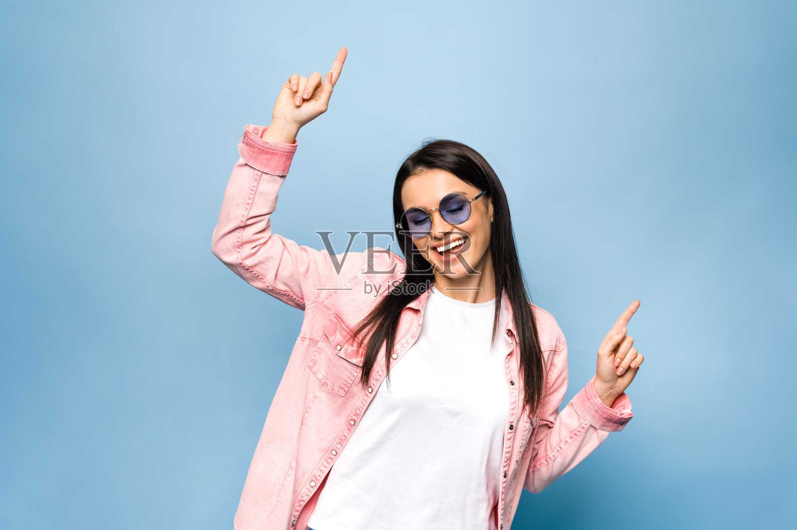 一个年轻迷人的开朗的白人女人的照片,穿着时尚的衣服和眼镜,跳舞和在孤立的蓝色背景上有乐趣照片摄影图片