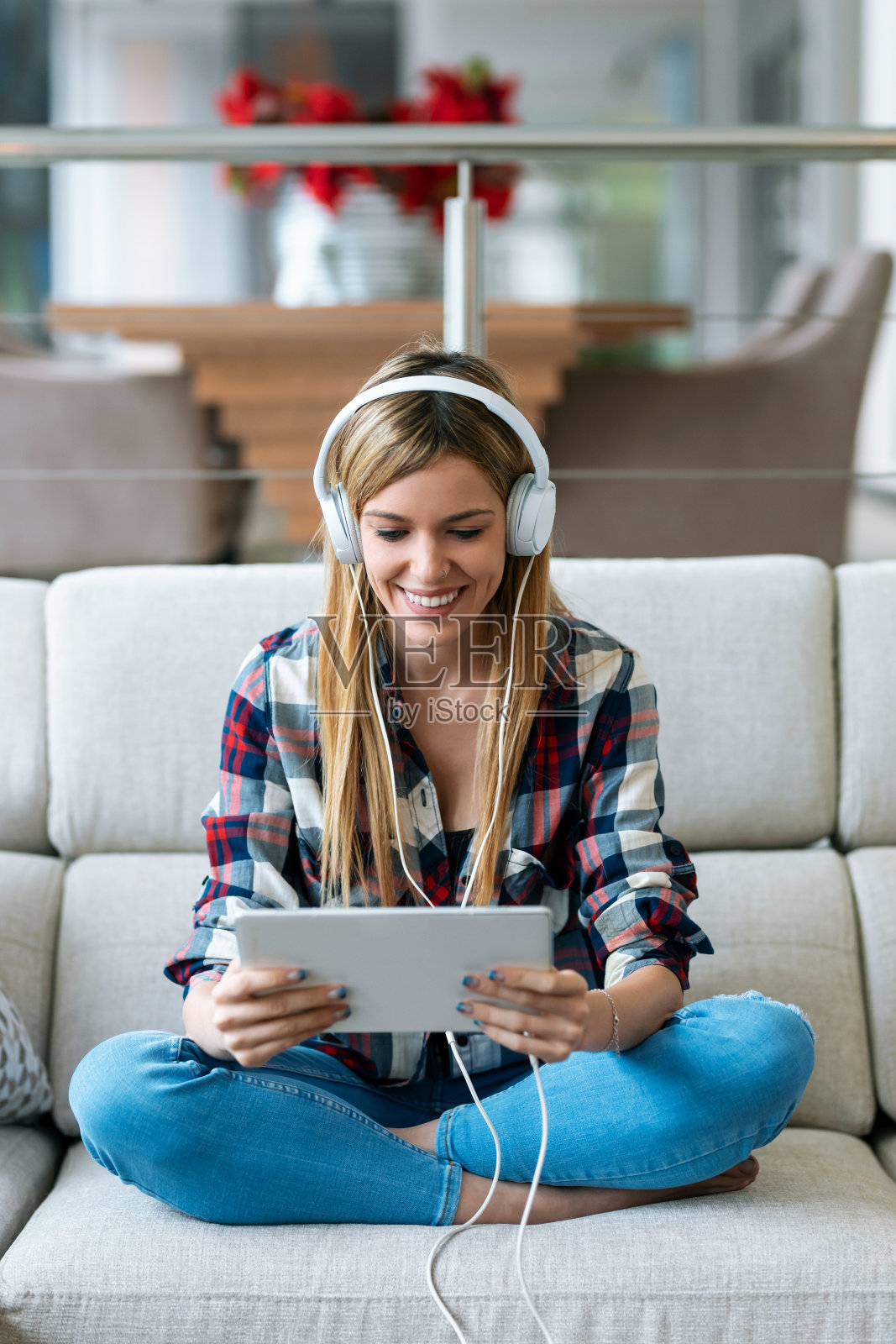 一个漂亮的年轻女子坐在家里的沙发上一边听着音乐一边用着她的数码平板电脑。照片摄影图片