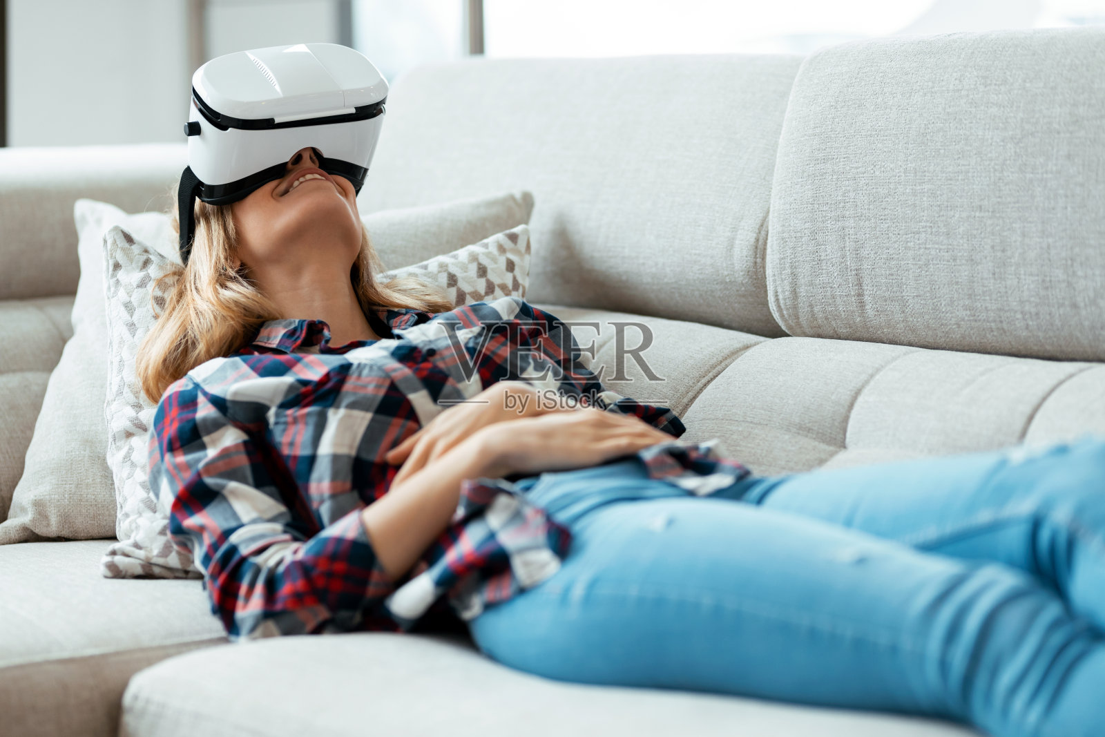 美丽的年轻女子使用虚拟现实头盔,享受此刻躺在家里的沙发上。照片摄影图片