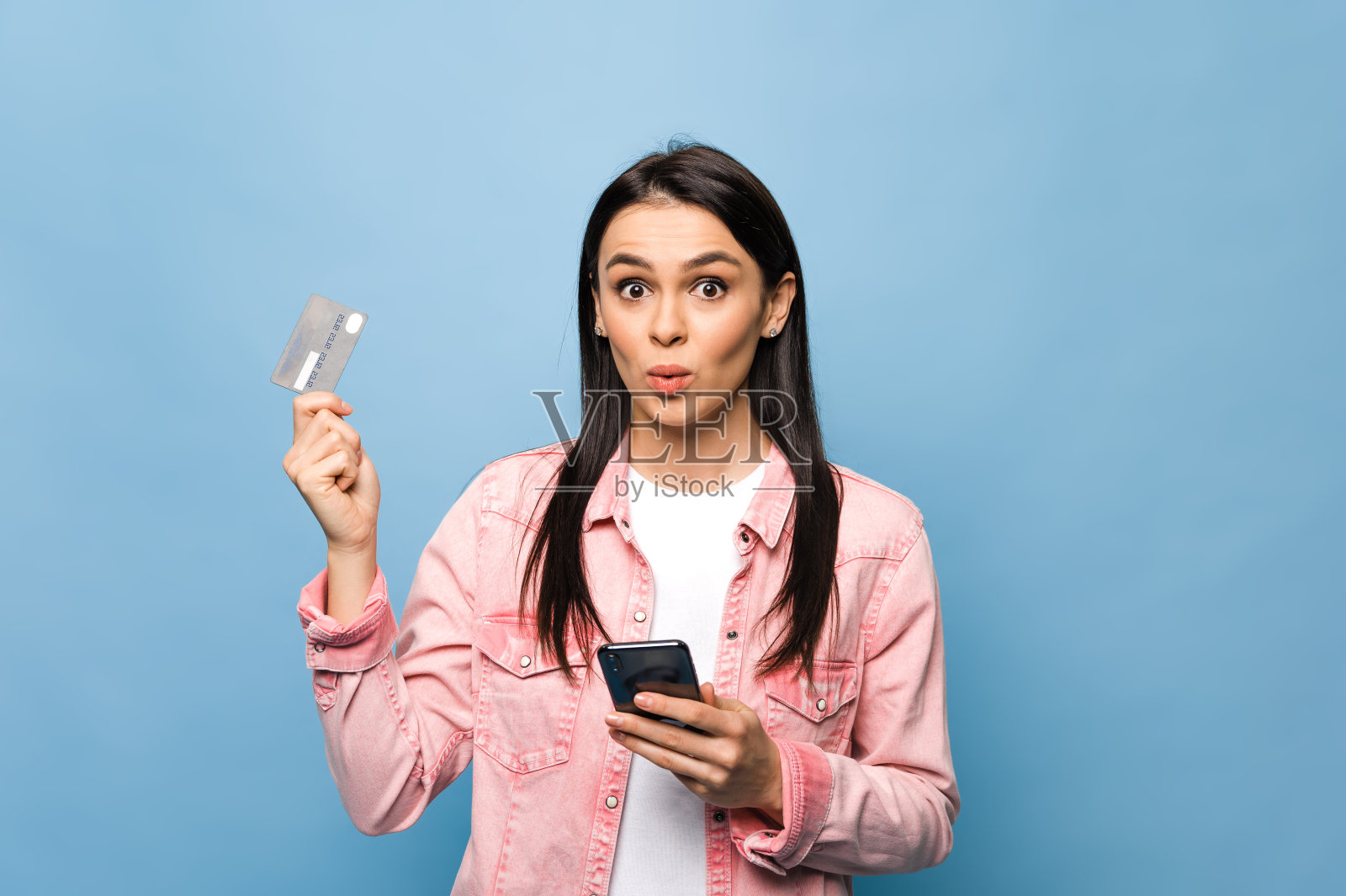 年轻的白人漂亮女人的肖像,用手机和银行信用卡购物或在线支付,站在孤立的蓝色背景。电子银行的概念照片摄影图片