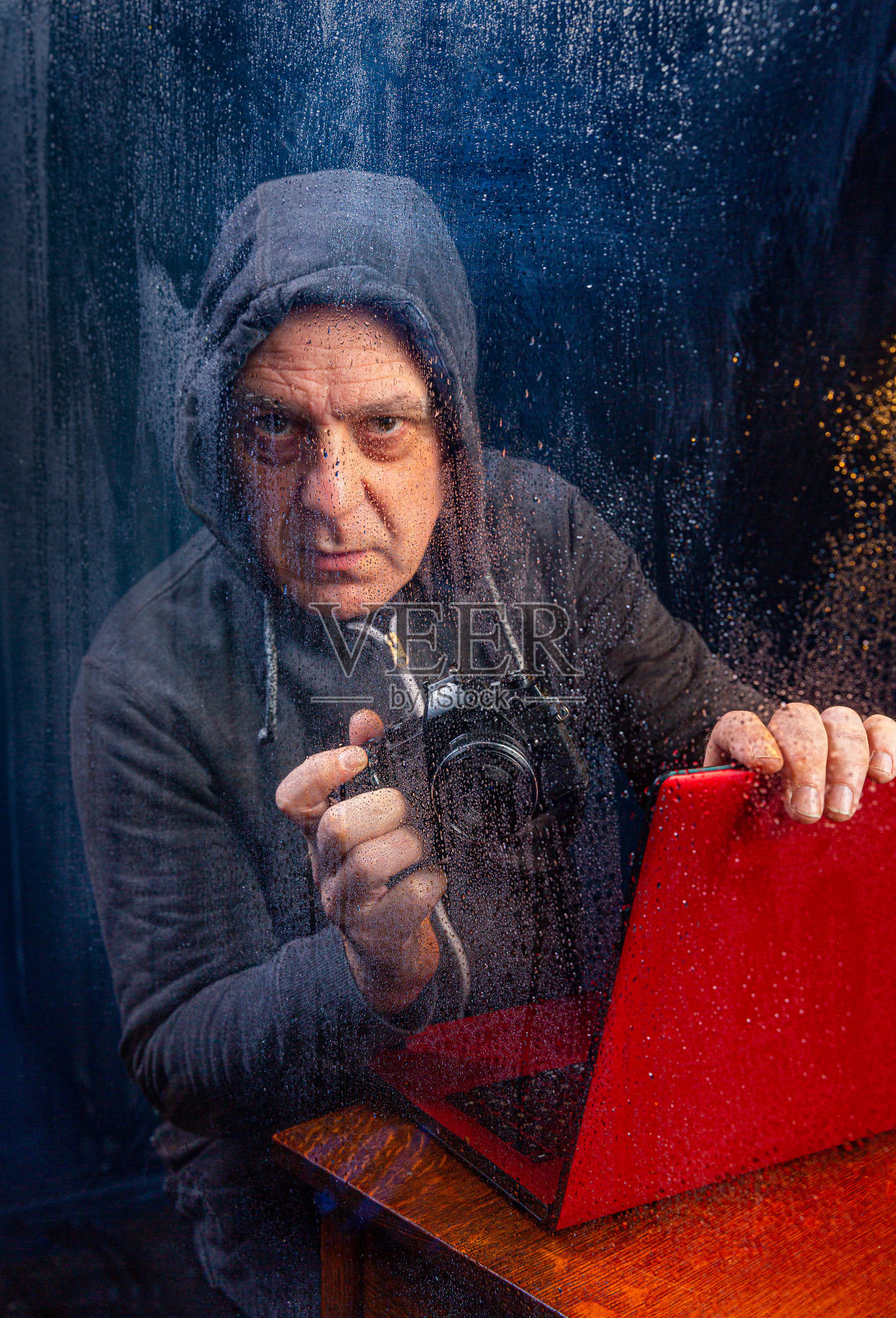 穿着帽衫的中年黑客,坐在下雨的窗户后面,带着相机和笔记本电脑,准备用勒索软件传播病毒照片摄影图片