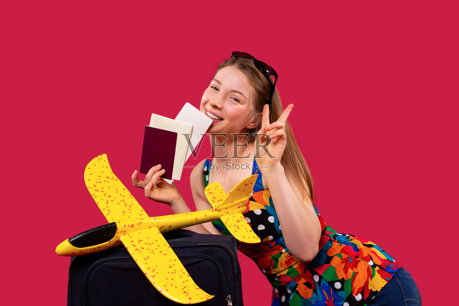 一个美丽的白种金发女郎对着镜头微笑,在旅行前拿着一个手提箱、文件和玩具飞机在红色背景下摆姿势。照片摄影图片