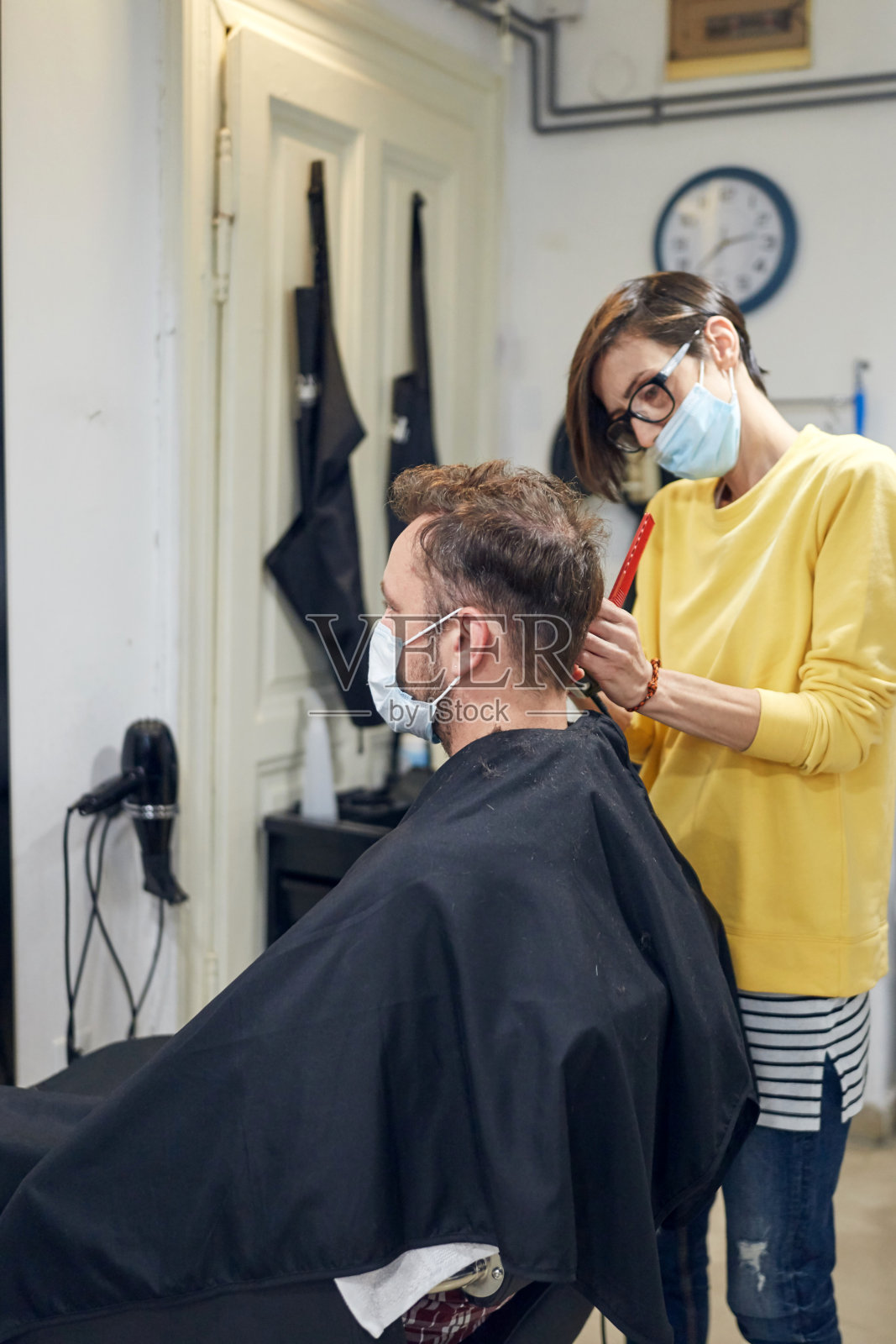 在一间沙龙里戴着医用口罩的理发师和顾客。戴着安全面罩工作。照片摄影图片