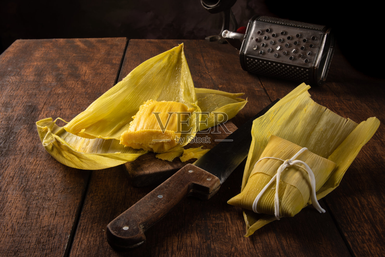 帕蒙哈,巴西玉米食品,叫帕蒙哈。在一张质朴的桌子上,放着一把小刀和一个旧的玉米磨碎器,低调的肖像,深色的抽象背景,选择性聚焦。照片摄影图片