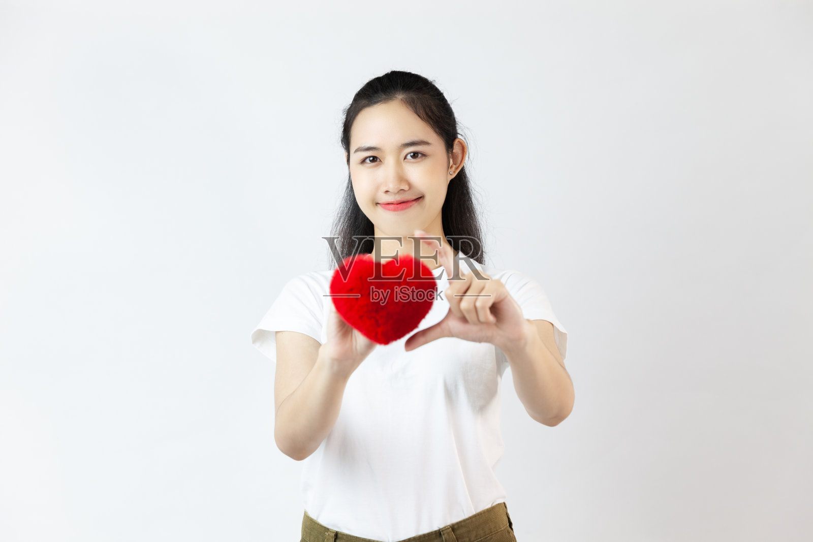 一个可爱的年轻亚洲女人穿着白色的t恤,拿着红色的心微笑着看着白色背景的相机。照片摄影图片