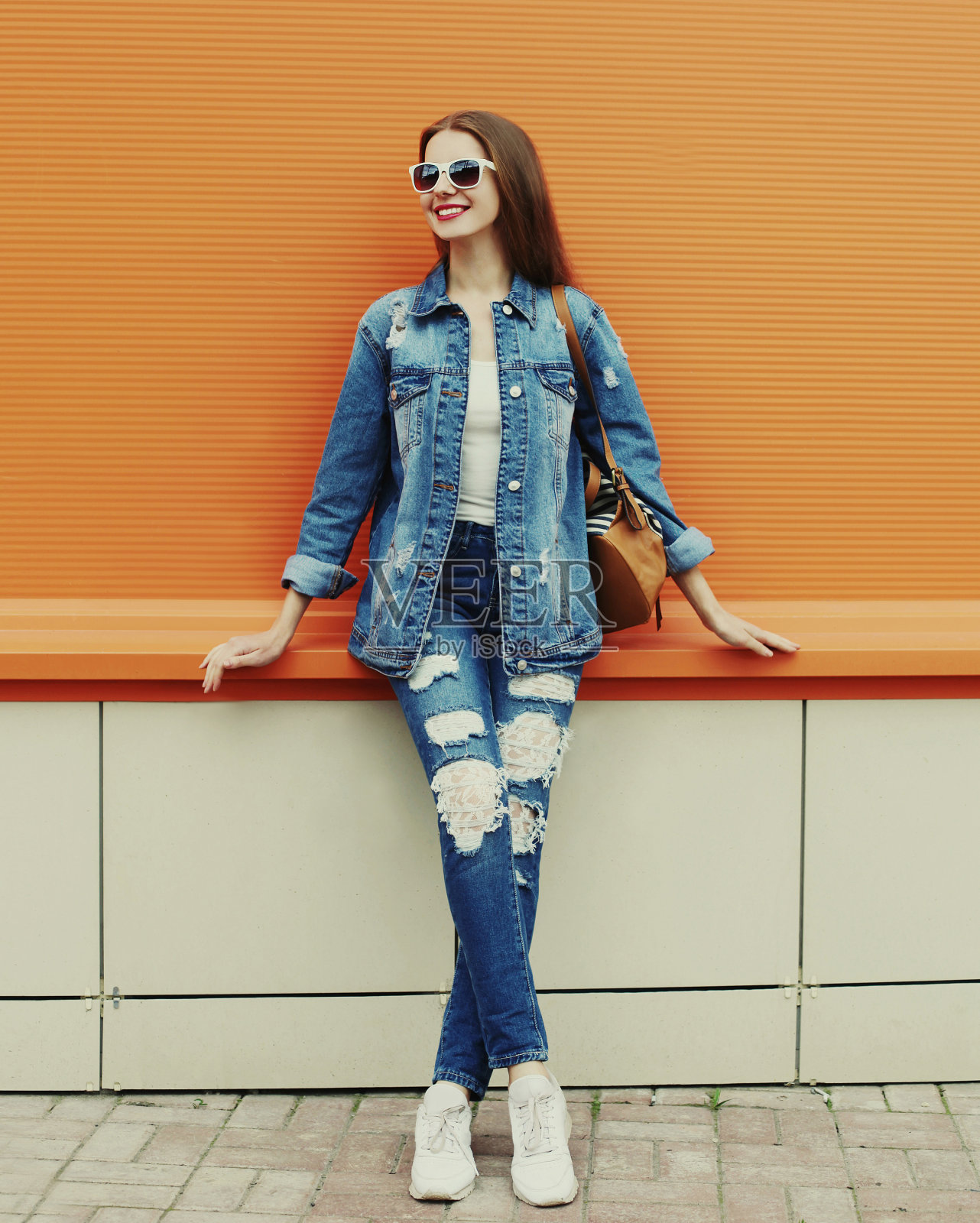 一个微笑的年轻女子的肖像,穿着牛仔夹克和牛仔裤,背包在一个橙色的背景照片摄影图片