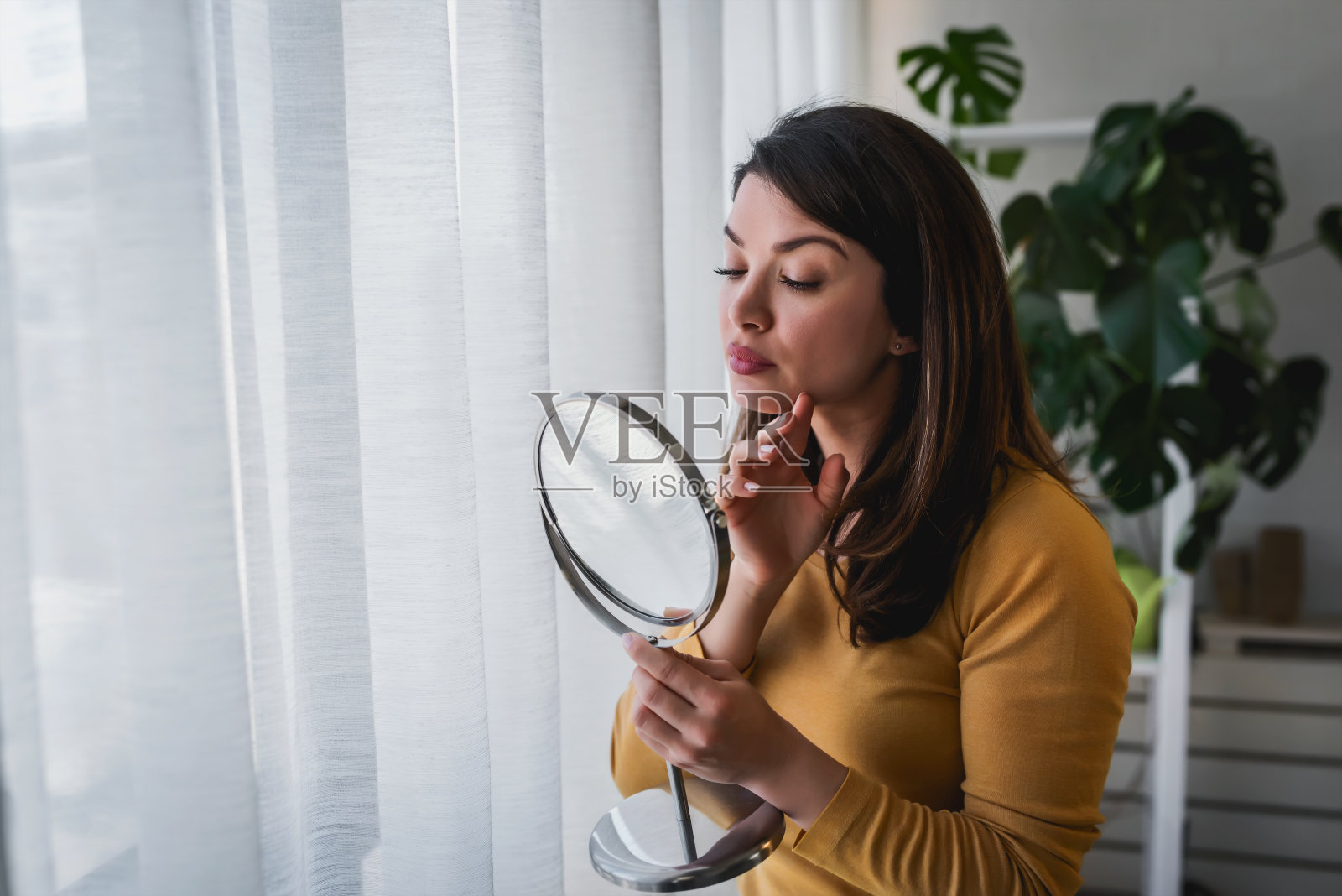 一个女人站在家里的大窗户边照镜子。她担心粉刺,面膜照片摄影图片