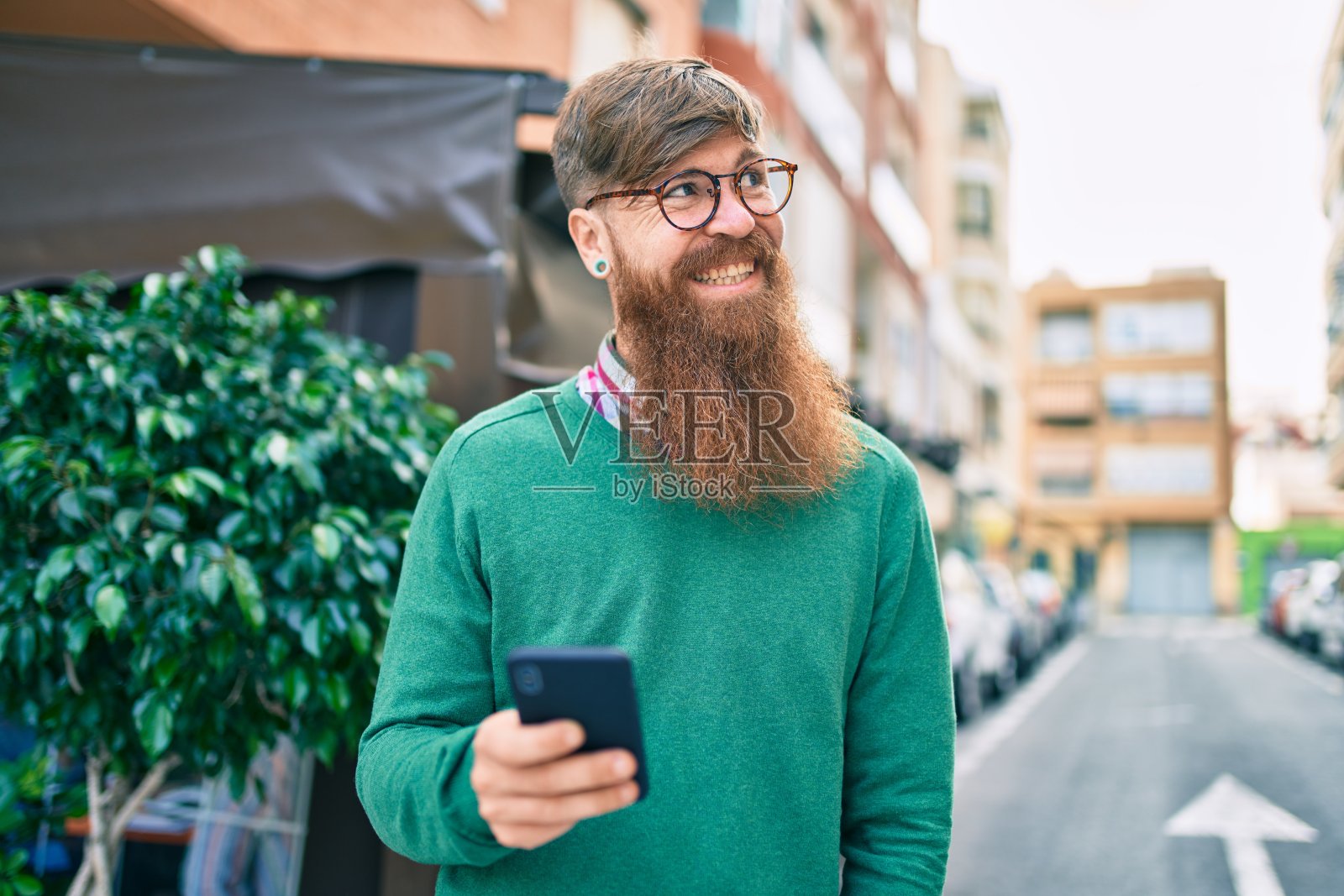 一个留着红胡子的年轻爱尔兰男子在城市里用智能手机微笑着。照片摄影图片