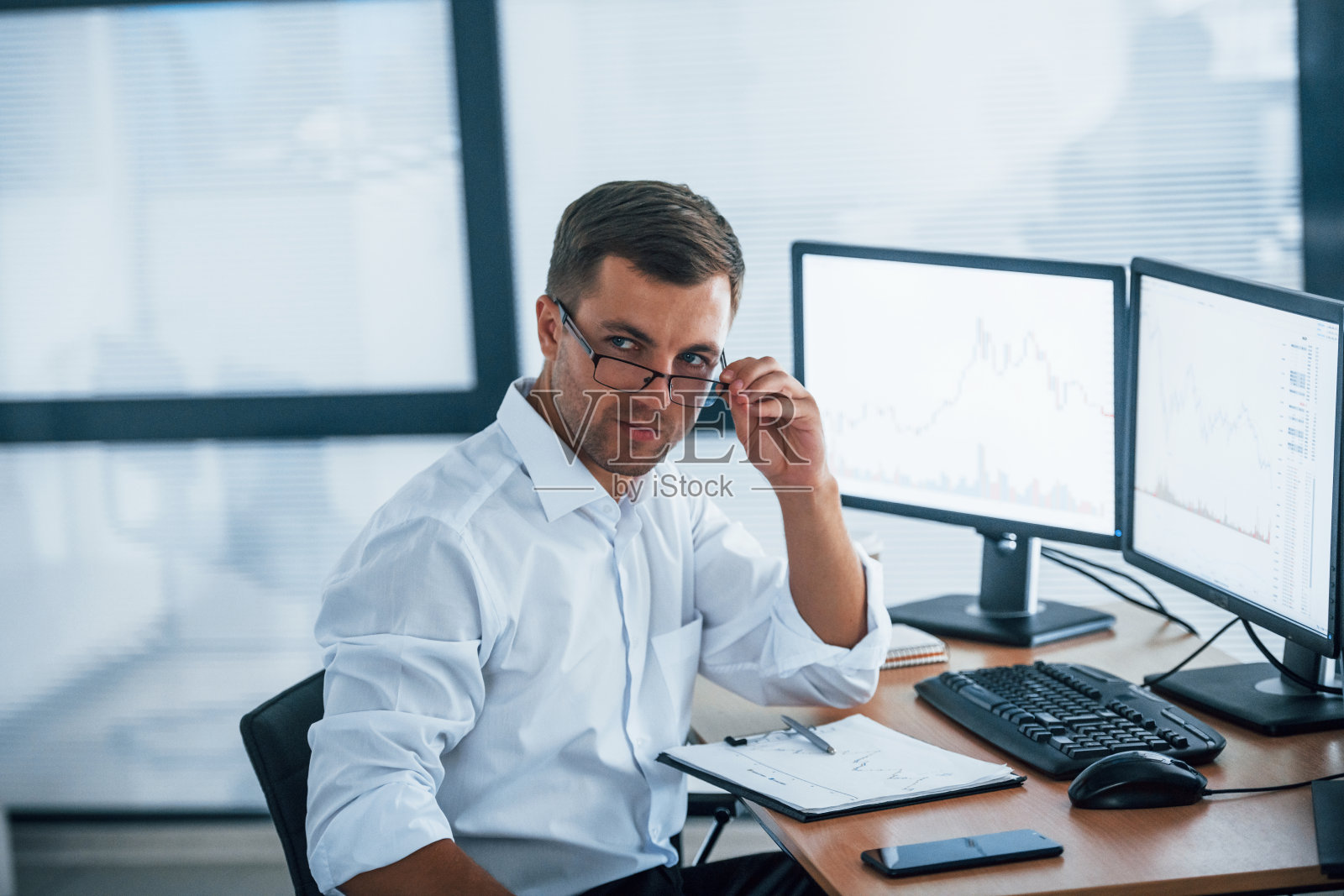 看着摄像机。穿着正装的年轻商人正在办公室里,办公室里有多个屏幕。交换和金钱的概念照片摄影图片