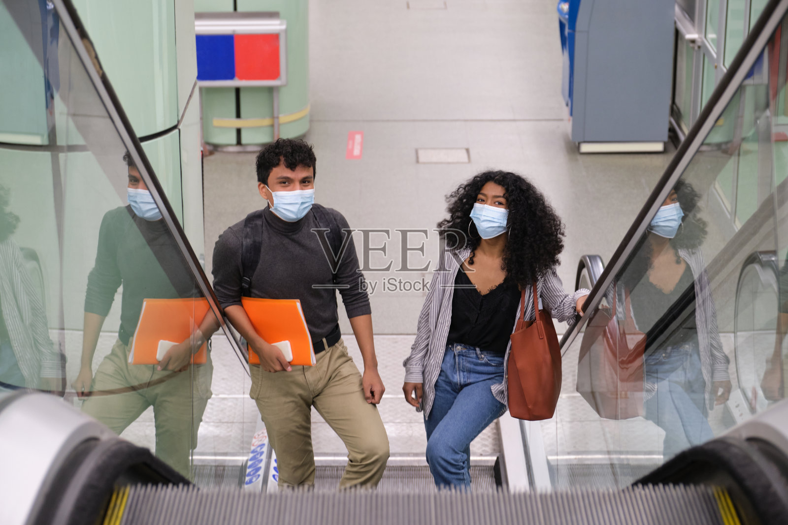 一对年轻的拉丁夫妇戴着防护面具走进火车站或地铁站的自动扶梯。公共交通的新常态。照片摄影图片