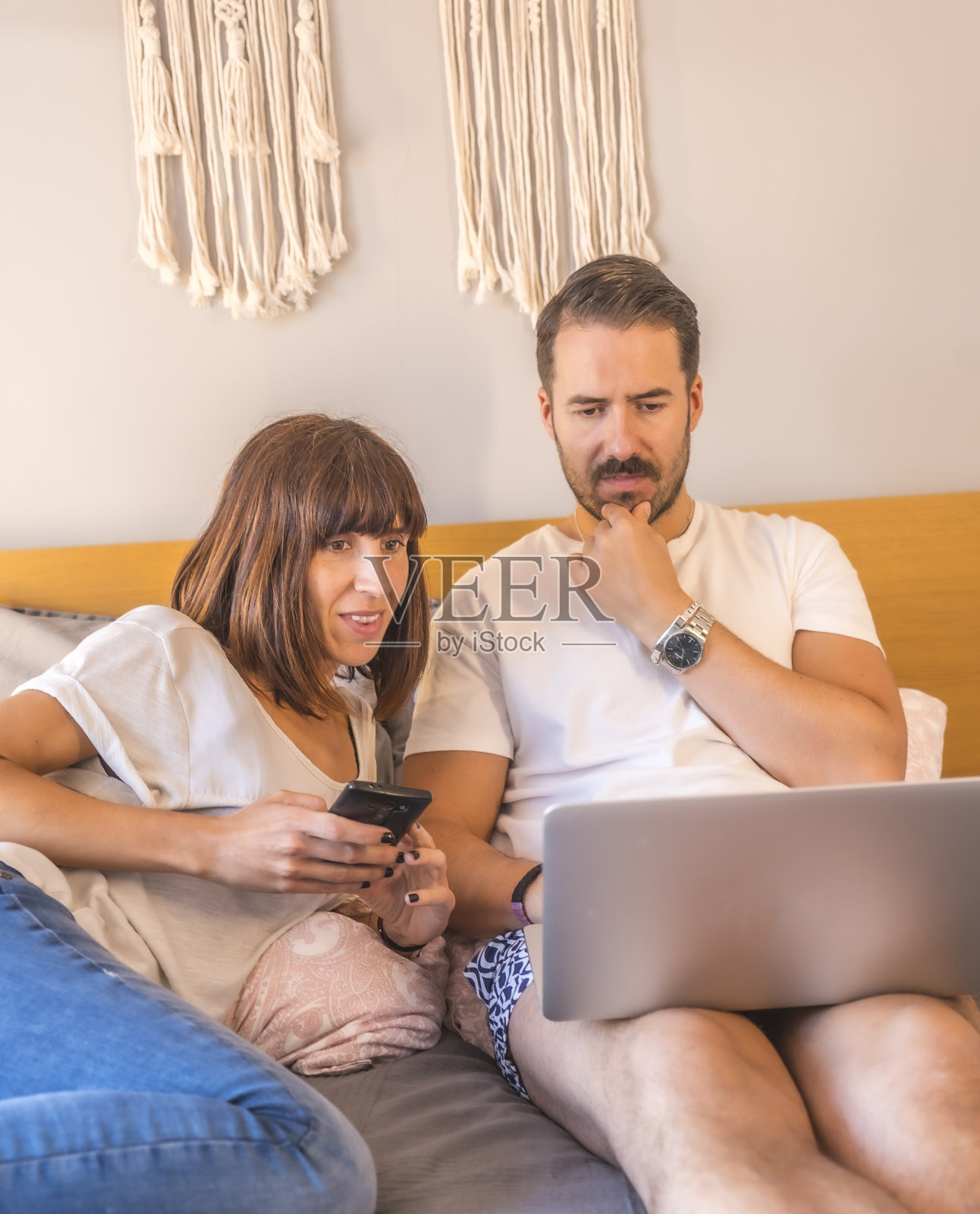 一对白人夫妇躺在床上,拿着电脑和电话,预订酒店或航班,安排假期,家庭使用新技术。照片摄影图片