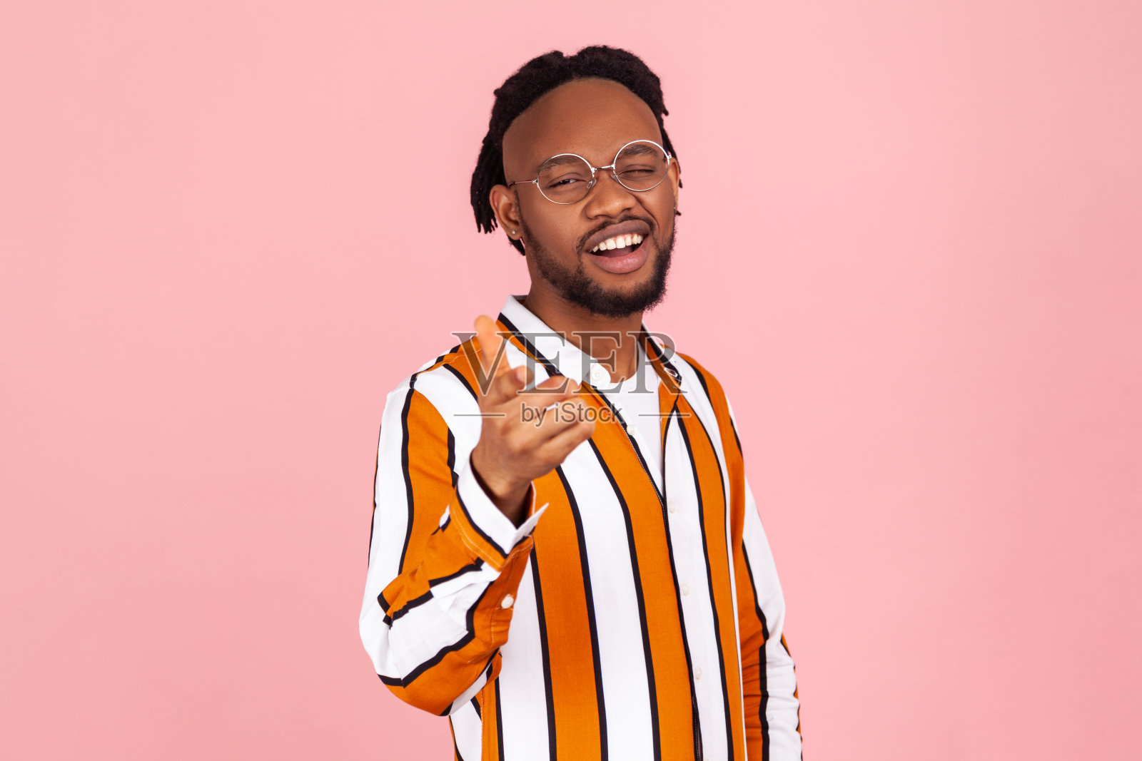 你过得如何?一个微笑着的美国黑人,留着胡子,梳着脏辫,戴着时髦的眼镜,穿着条纹衬衫,指着你眨着眼睛照片摄影图片