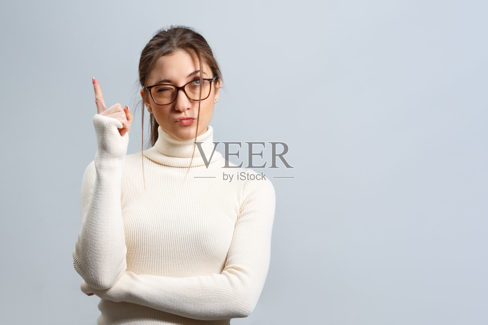 戴着眼镜的美丽年轻女孩沉思着,她在一个孤立的灰色背景上举起了手指。照片摄影图片
