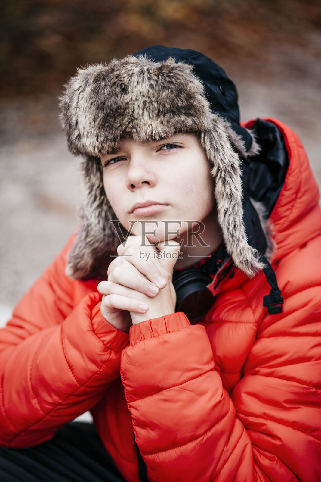 帅气聪明的少年坐着,看着镜头,手放在下巴上,看起来严肃,自信,富有挑战性,聪明,紧张,戴着毛皮俄罗斯帽,红色夹克-近距离,肖像,大头照。照片摄影图片