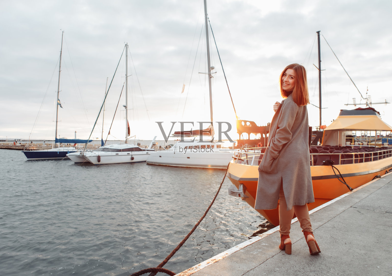 日出时分,一个穿着秋日大衣的年轻快乐的女人在游艇俱乐部摆姿势照片摄影图片