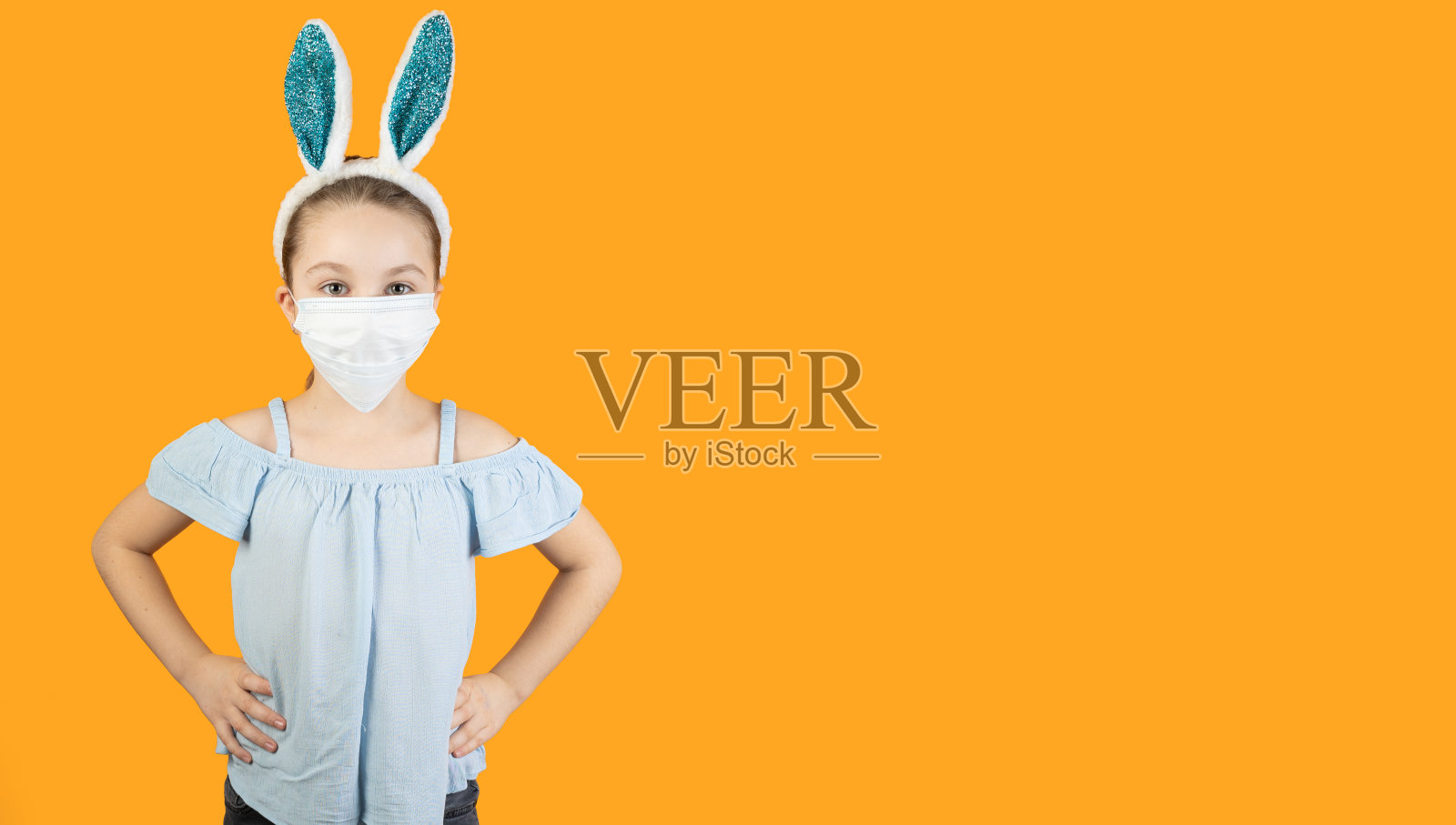 一个小女孩戴着冠状病毒的医用口罩,头戴兔耳,双手放在臀部。复活节和春天的象征。照片摄影图片