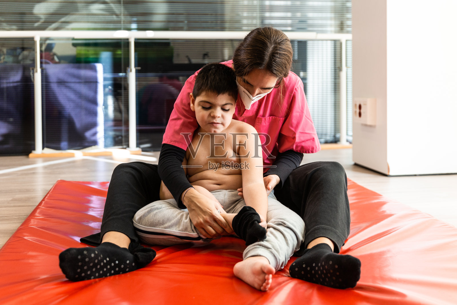 残疾儿童和理疗师在红色体操垫上做练习。大流行面具保护照片摄影图片