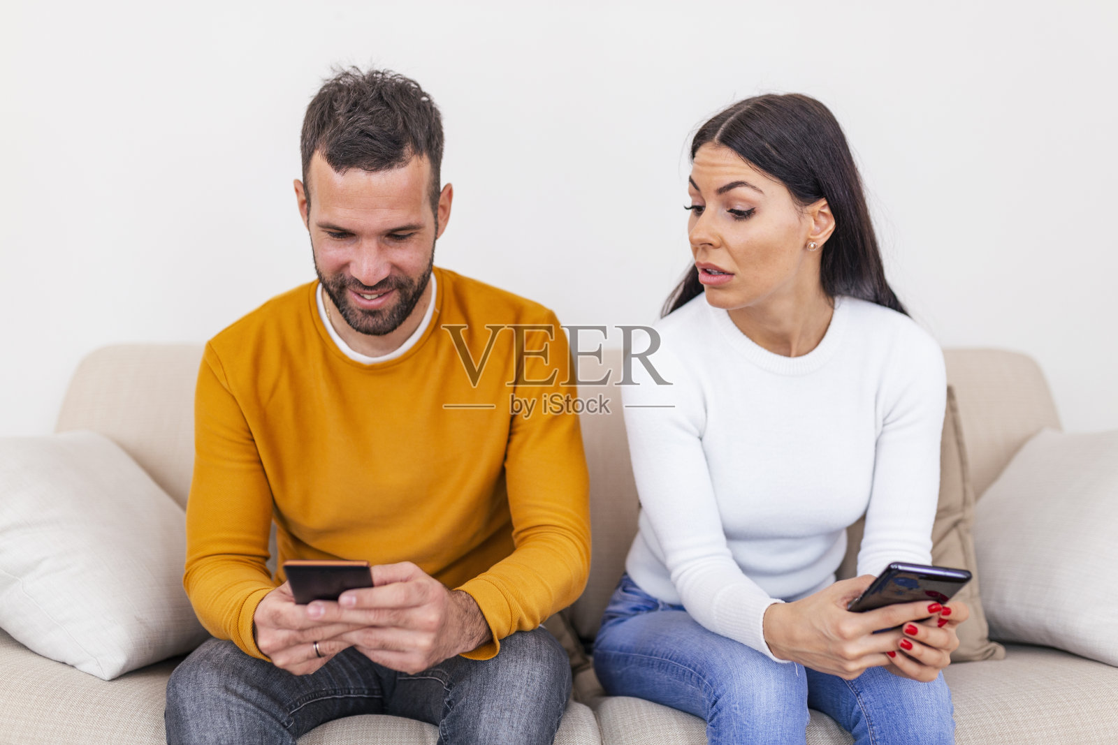 嫉妒的年轻女子用智能手机看着微笑的男友在家里使用智能手机,关系问题概念。不信任的概念照片摄影图片