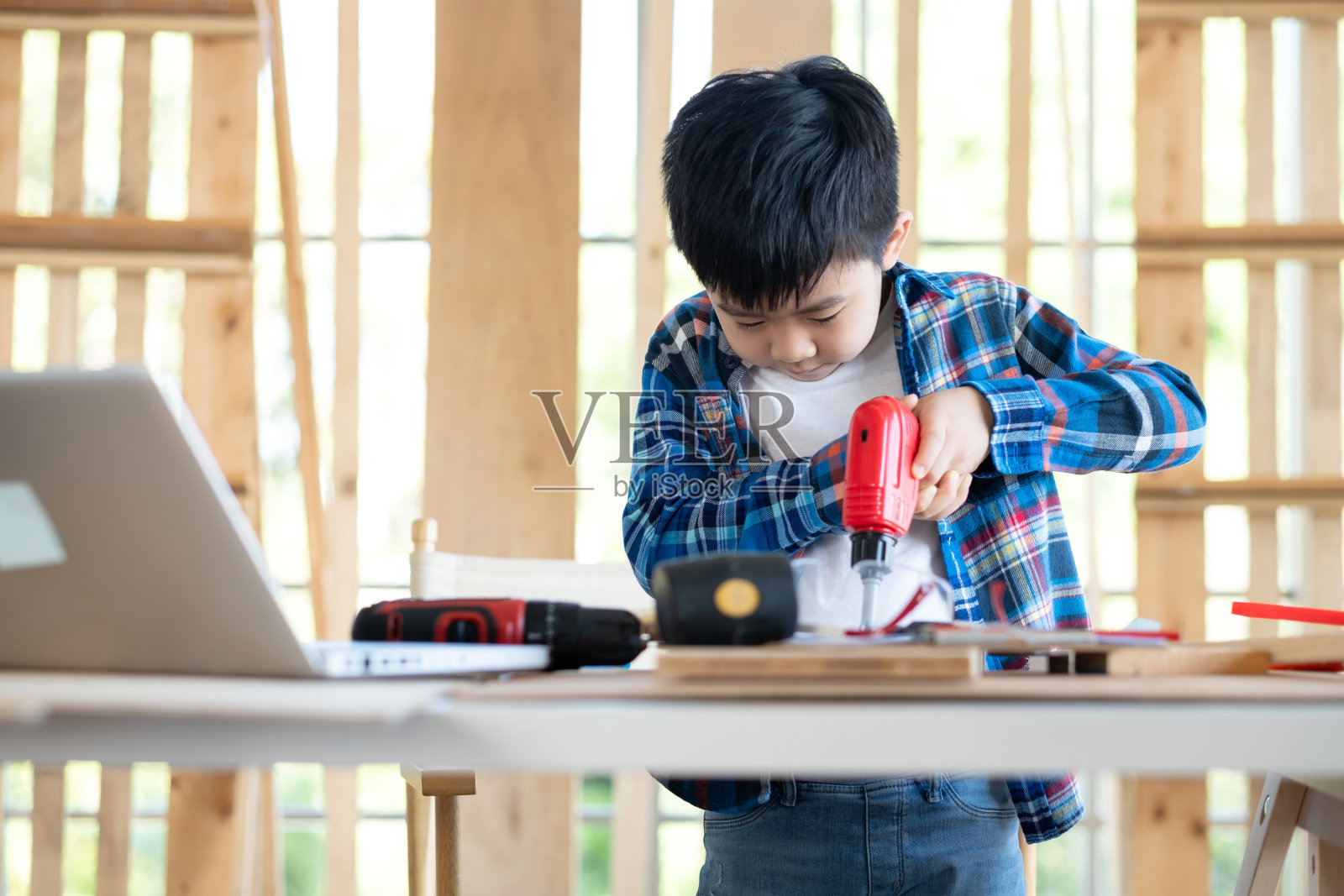 一个小男孩穿着衬衫,用螺丝刀试着螺丝在教室里的木片。桌子上有笔记本电脑和许多设备。教育、学前班和学习理念。照片摄影图片