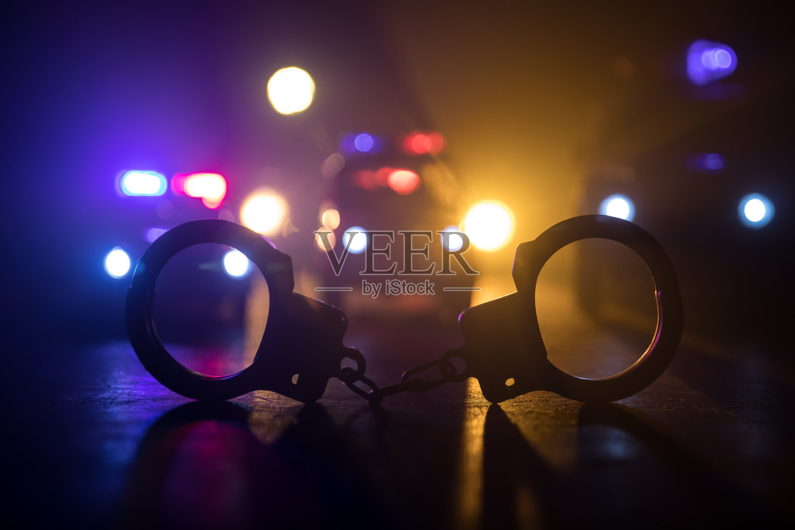 警方夜间突袭,你将被逮捕。手铐的轮廓和警车在后面。在雾蒙蒙的背景下,红色和蓝色警灯闪烁。照片摄影图片