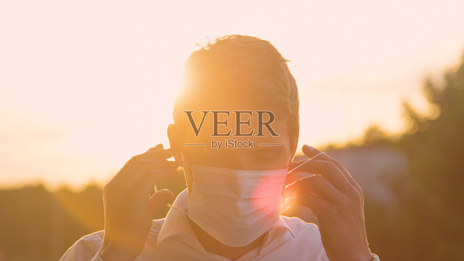 特写:在金色的夕阳下,微笑的白人男子戴上了一次性口罩。照片摄影图片