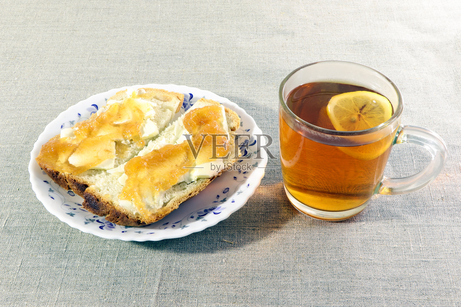 两片涂着黄油和果酱的白面包放在盘子里。一杯柠檬茶。明亮的背景。早餐。照片摄影图片