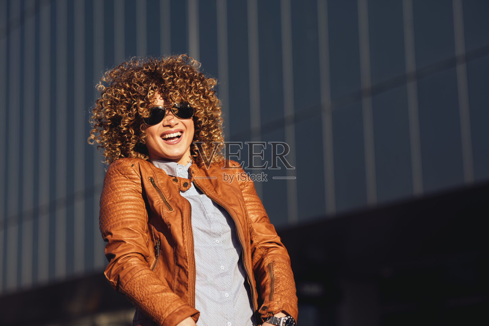一个年轻快乐的女人的肖像,穿着棕色皮夹克,金色的非洲式卷发,戴着太阳镜在街上照片摄影图片