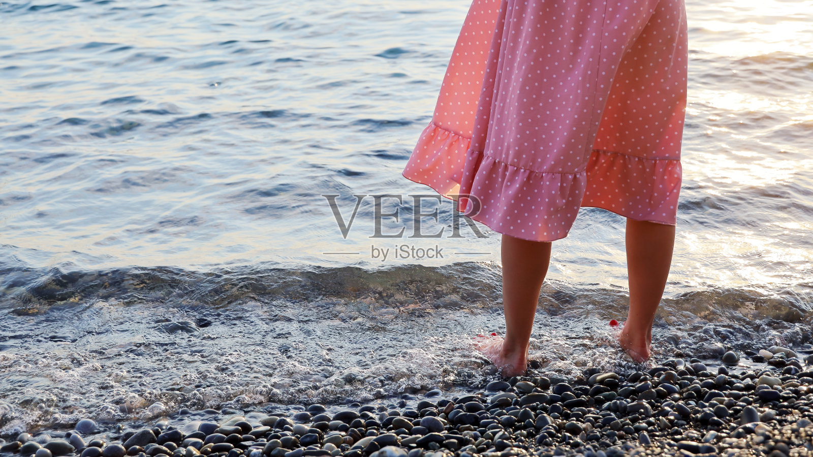穿着粉红色裙子的女人的腿赤脚站在海水卵石岸边照片摄影图片