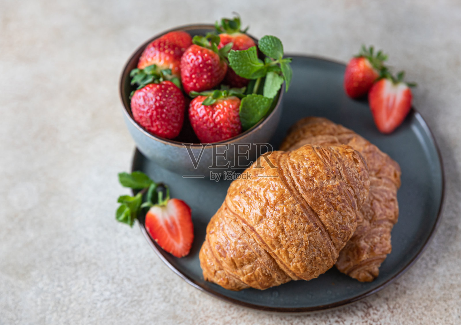 法式脆皮牛角面包,草莓盘,浆果碗,混凝土背景。新鲜的烤羊角面包。早餐或甜点的主意。照片摄影图片