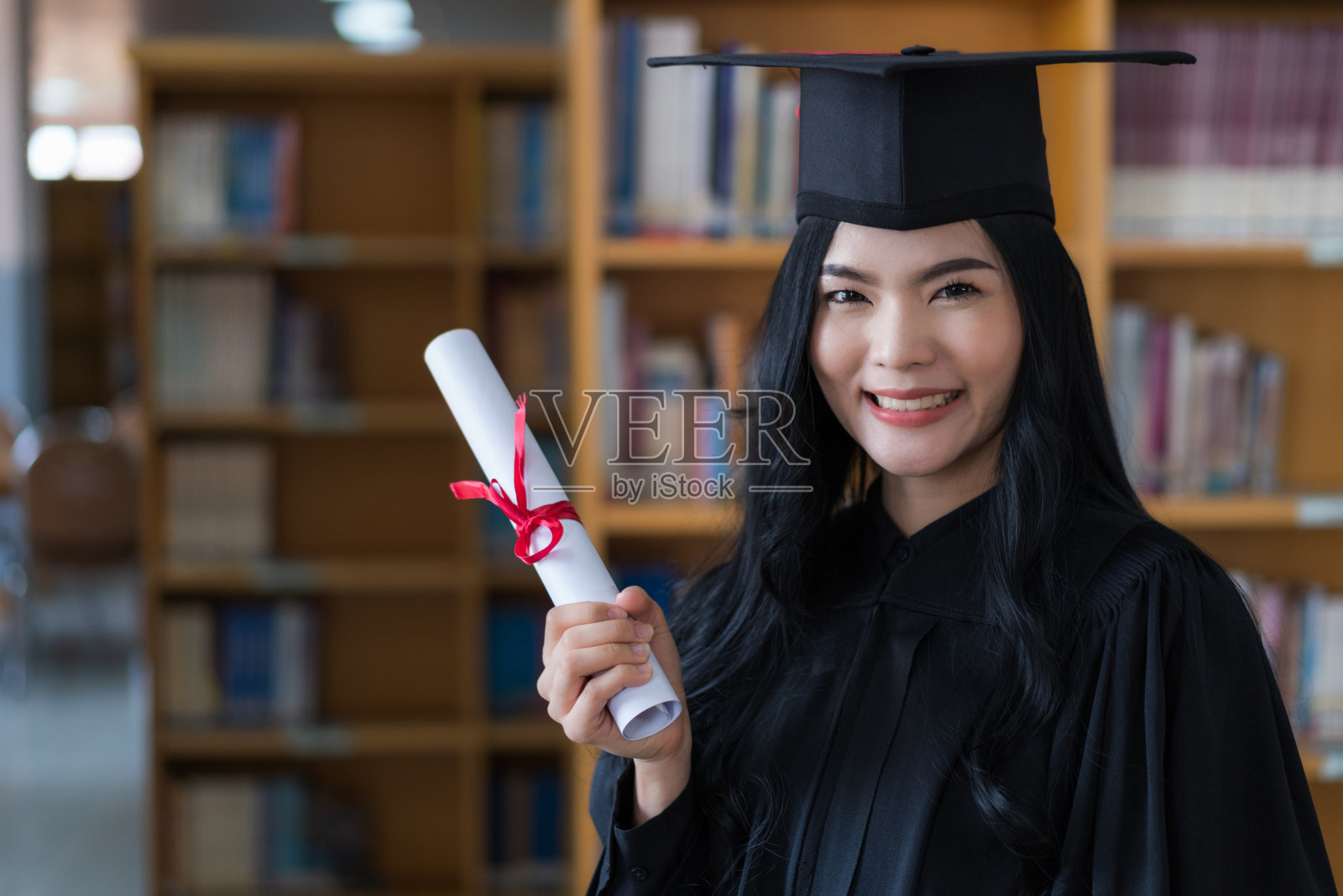 一名年轻快乐的亚洲女子大学毕业生穿着毕业礼服和帽子戴着面具持有学位证书,以庆祝她的教育成就在毕业典礼上。库存图片照片摄影图片