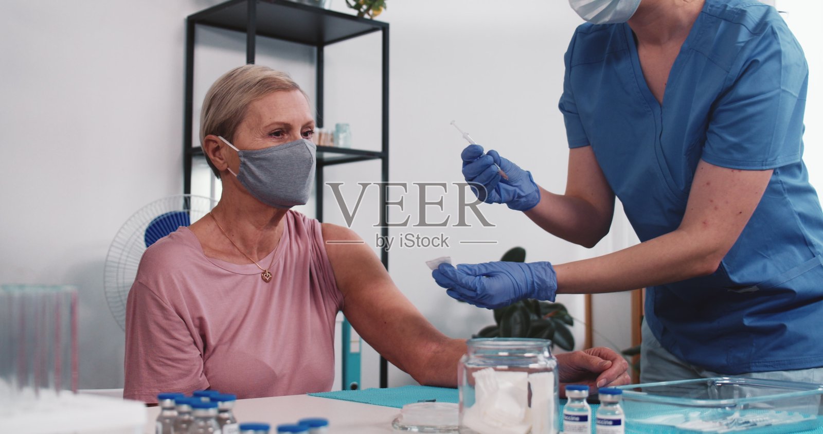 美丽的资深白种人妇女接种了年轻医生在蓝色的手术服,接受注射在轻诊所照片摄影图片